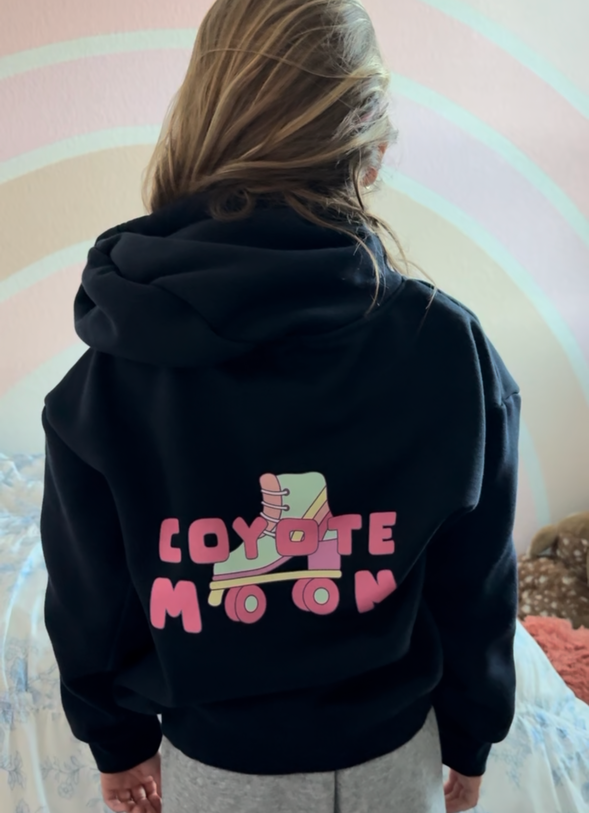 Person wearing a black hoodie with 'Coyote Moon' design on the back, standing in a room with a pink and white circular pattern.